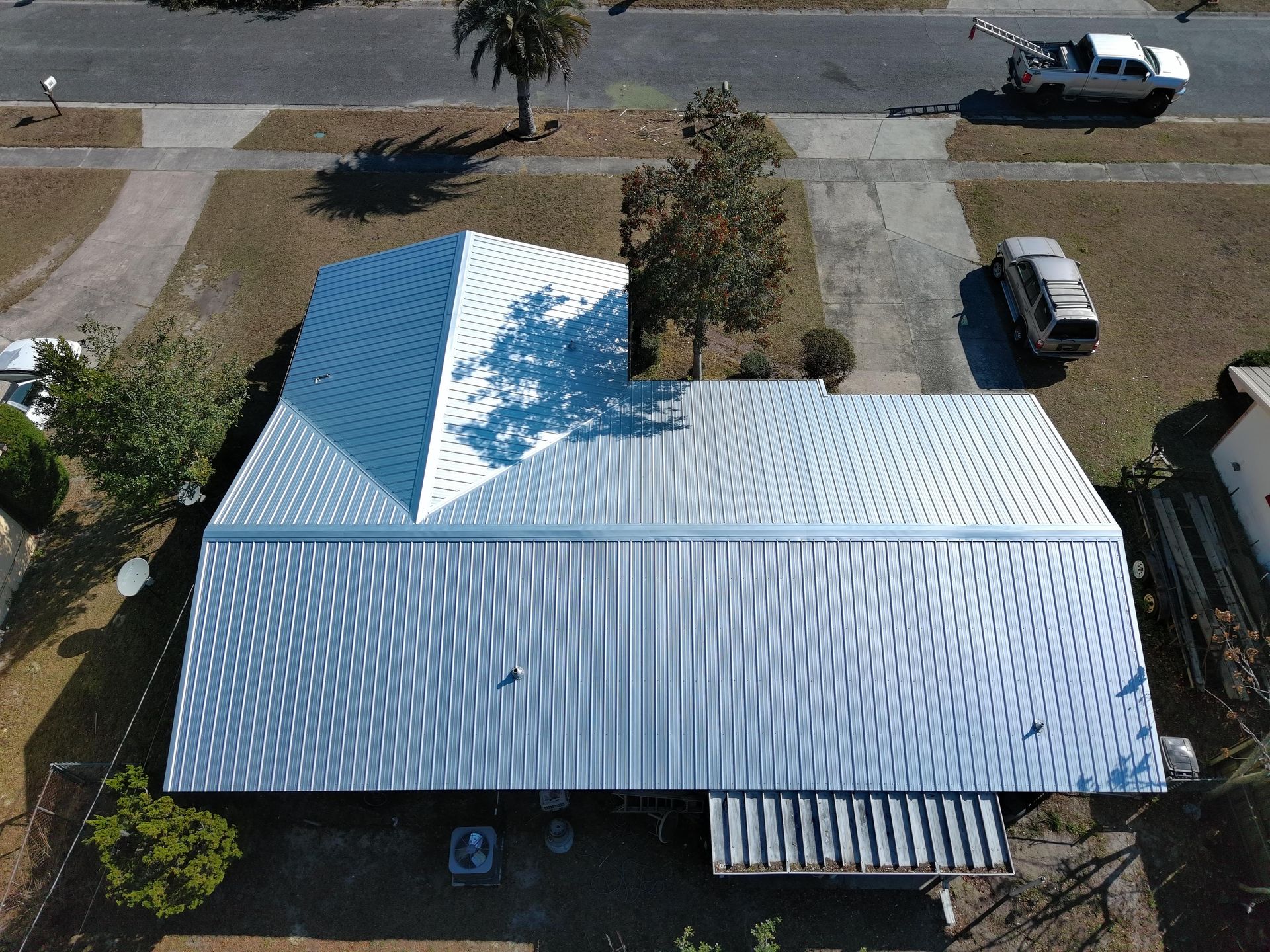 An aerial view of a silver, patterned metal roof on a house with nearby vehicles, trees, and a driveway.