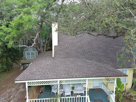A brown single-story house with a gray roof and a tan garage door sits behind a gravel driveway under a clear blue sky.