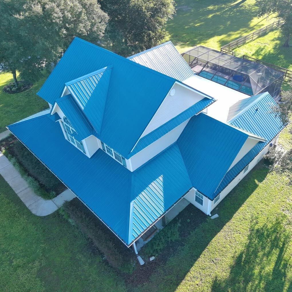 Aerial view of a suburban house with a bright blue metal roof, white siding, and a backyard pool enclosure.