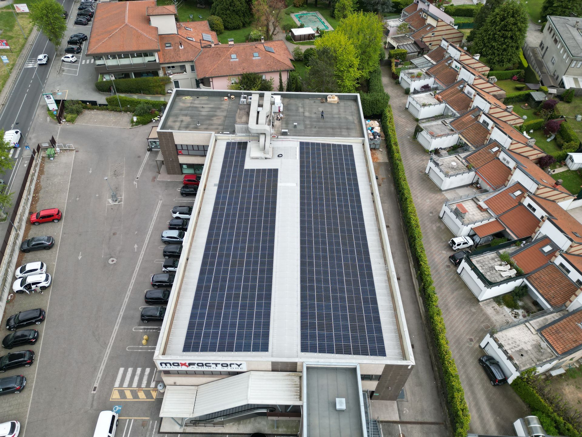 Vista aerea di un edificio con pannelli solari sul tetto. Auto parcheggiate di fronte, case residenziali sulla destra.