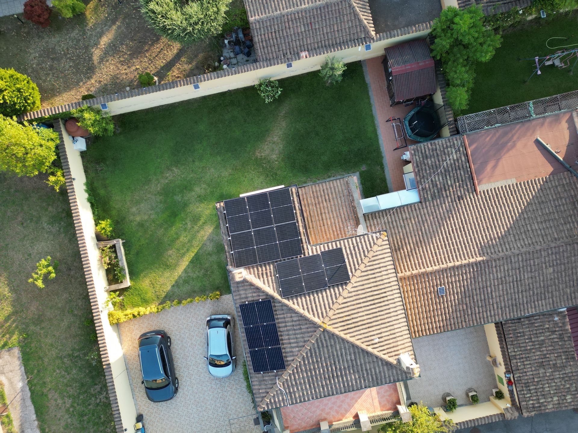 Vista aerea di una casa con pannelli solari sul tetto, un cortile e due auto parcheggiate.
