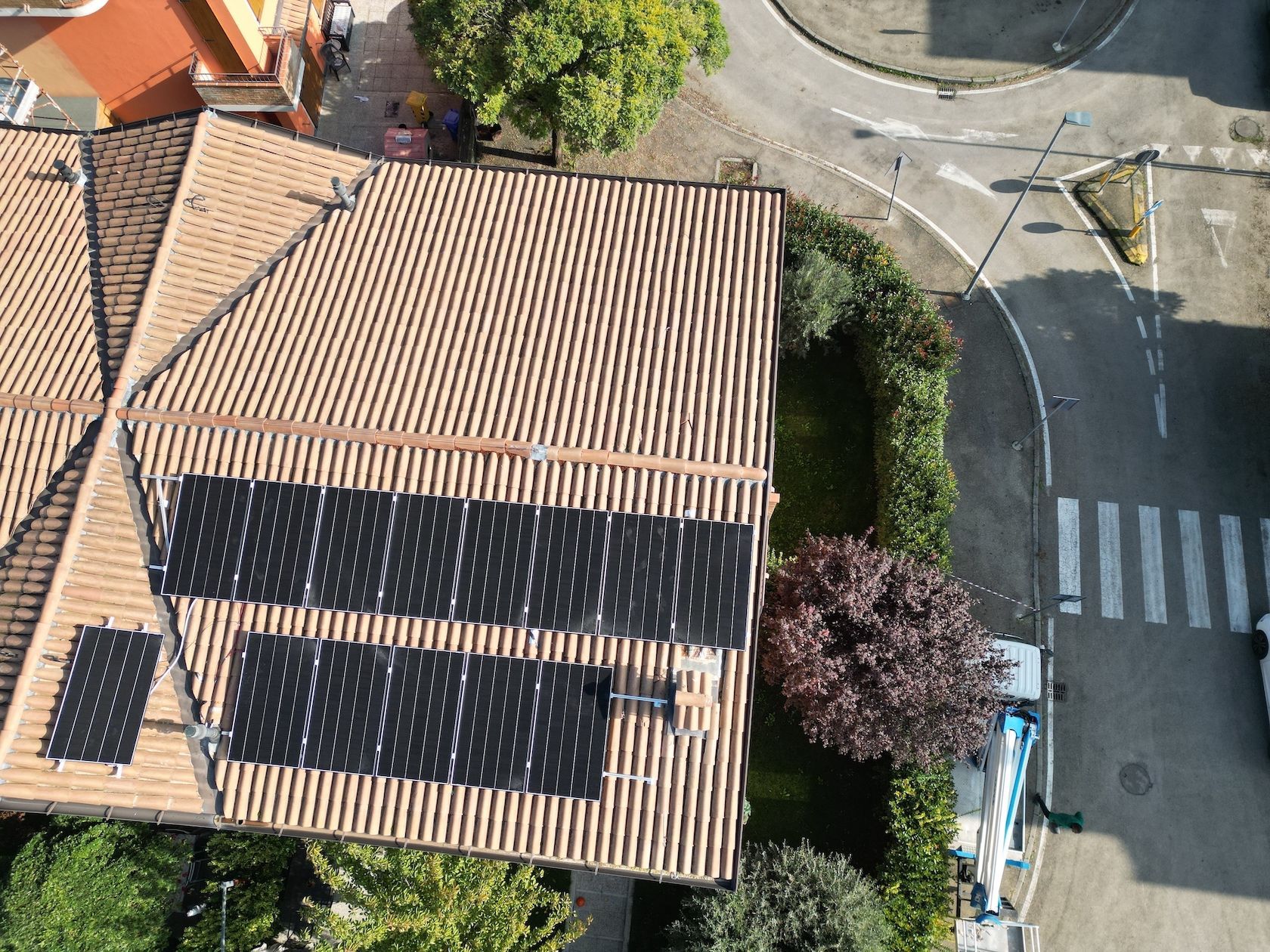 Vista dall'alto di una casa con pannelli solari su un tetto di tegole marroni, vicino a una strada con attraversamenti pedonali e alberi.