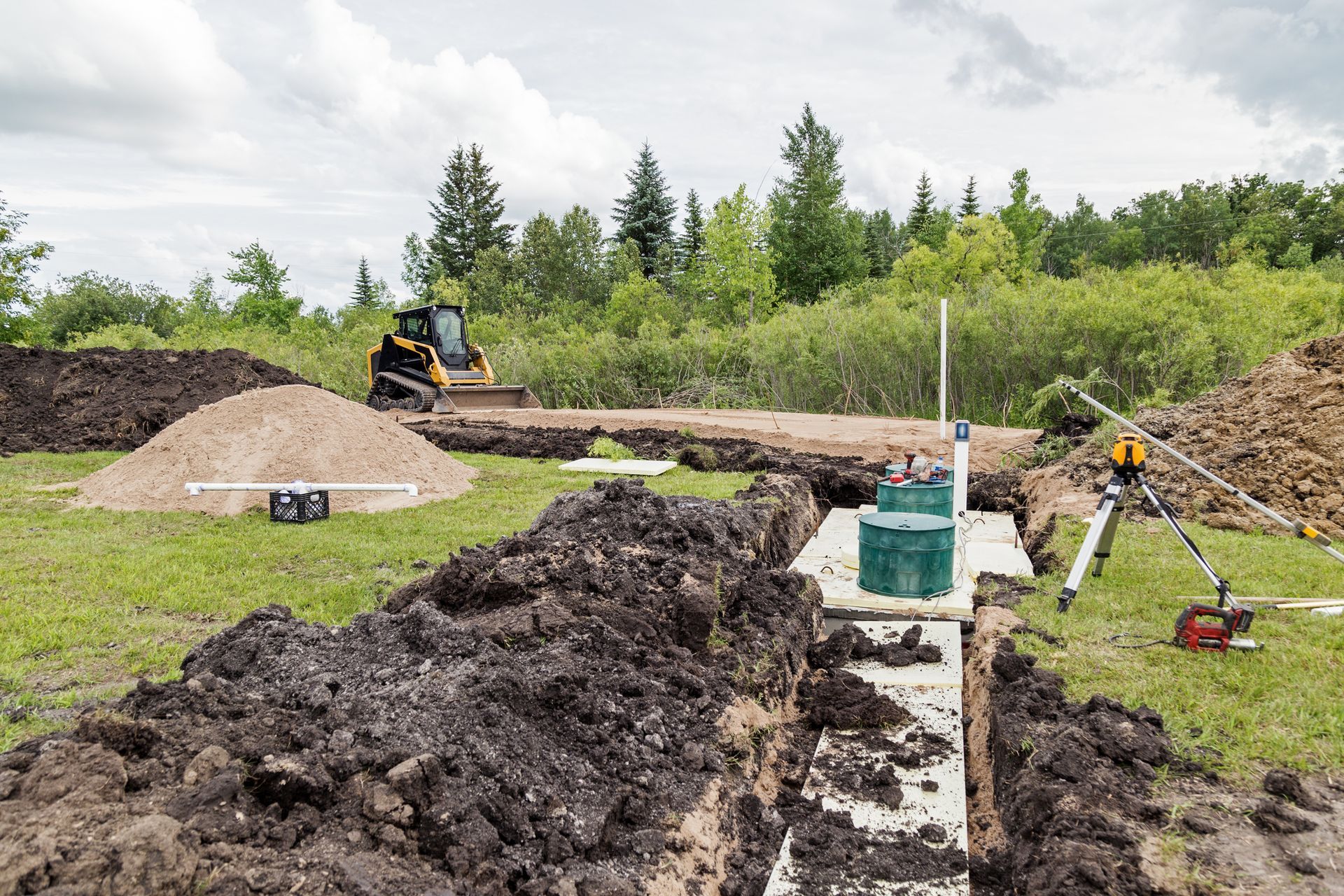 Construction site with trenches, a small excavator, and septic tank installation. Outdoor setting with dirt piles.