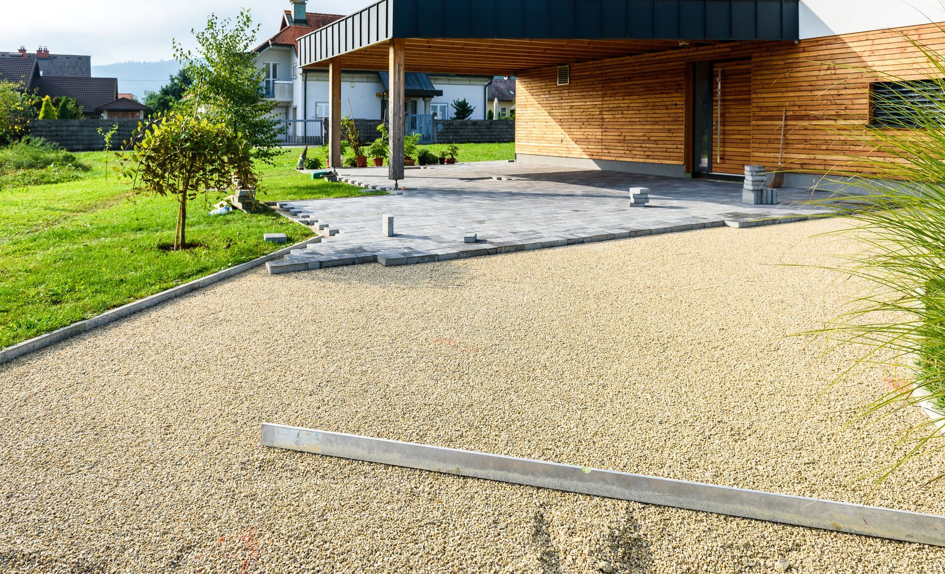 Driveway with light-colored gravel leading to a modern carport; green lawn with a tree and house in background.