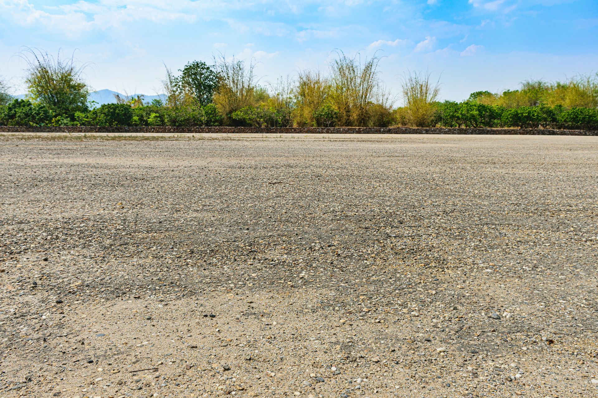 Gravel road stretches towards a line of trees against a blue sky with sparse clouds.