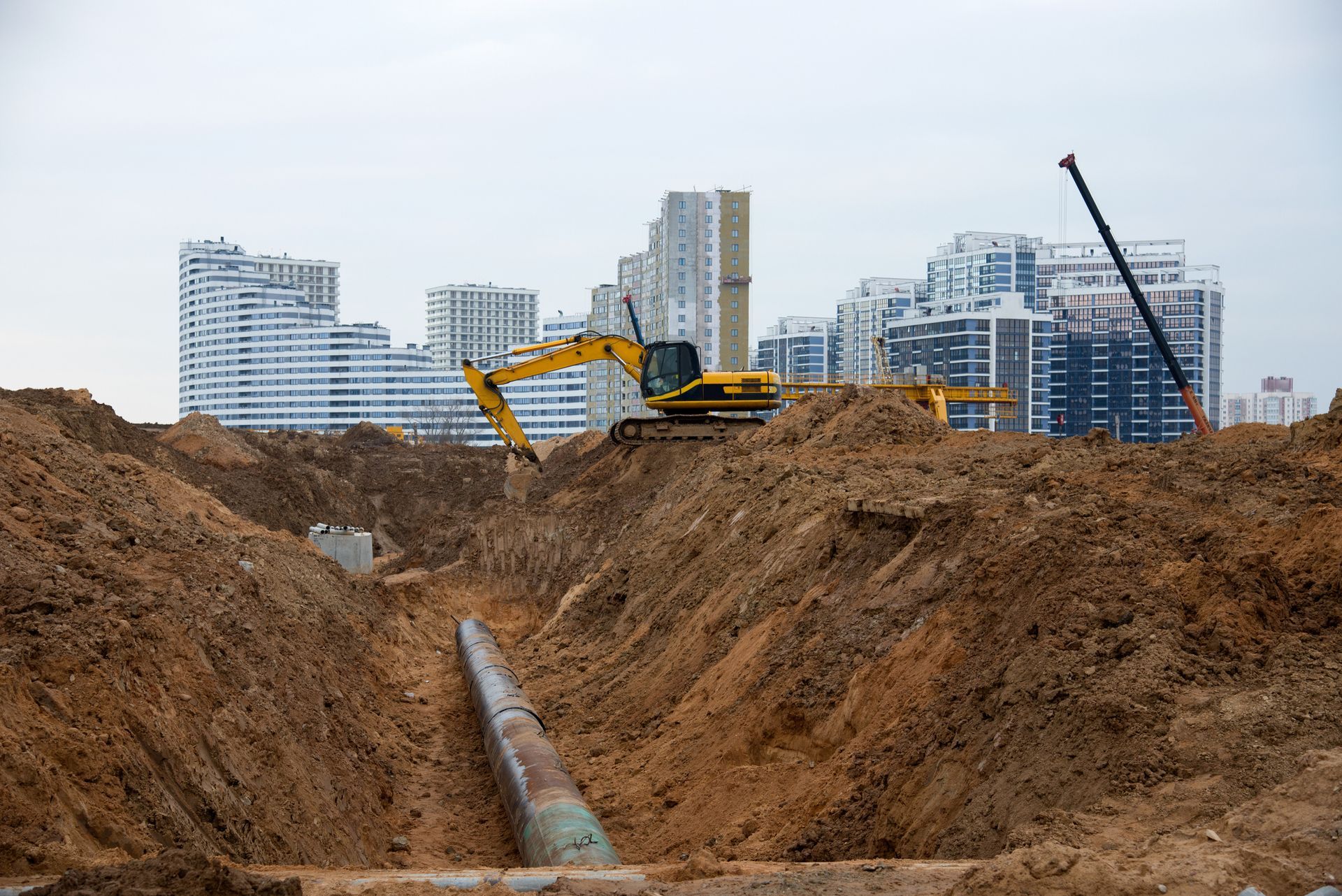 Excavator working on a pipeline in a trench; apartment buildings in the background under a cloudy sky.