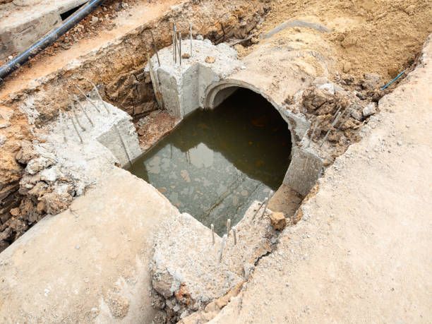 Open sewer pipes in a dirt trench, surrounded by concrete. Water fills the open area.
