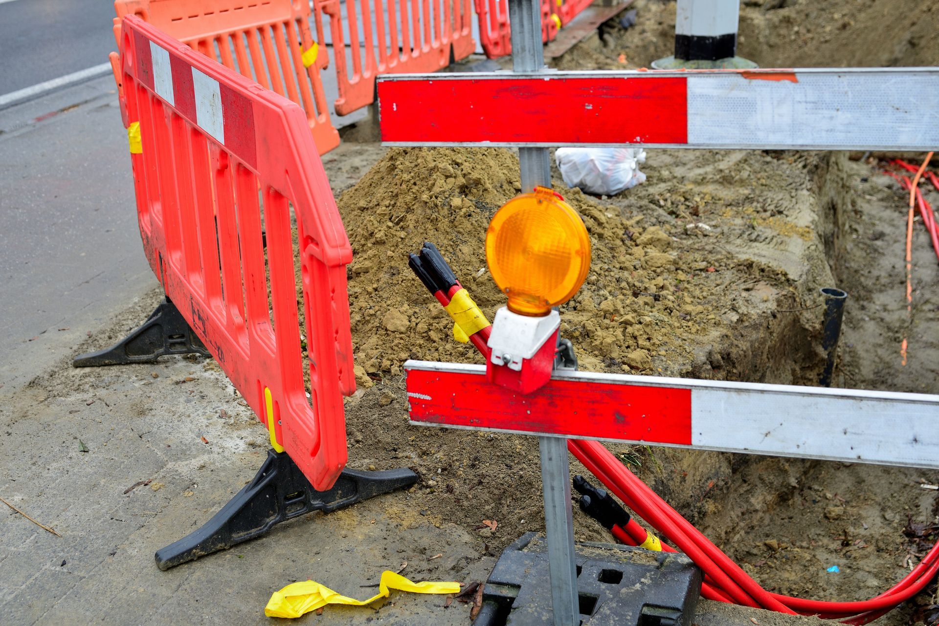 Orange and white traffic barriers surround an open construction trench. A flashing amber light is attached.