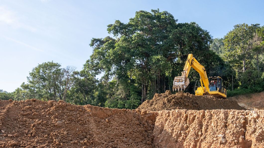 Yellow excavator digging into a dirt mound next to a forest.