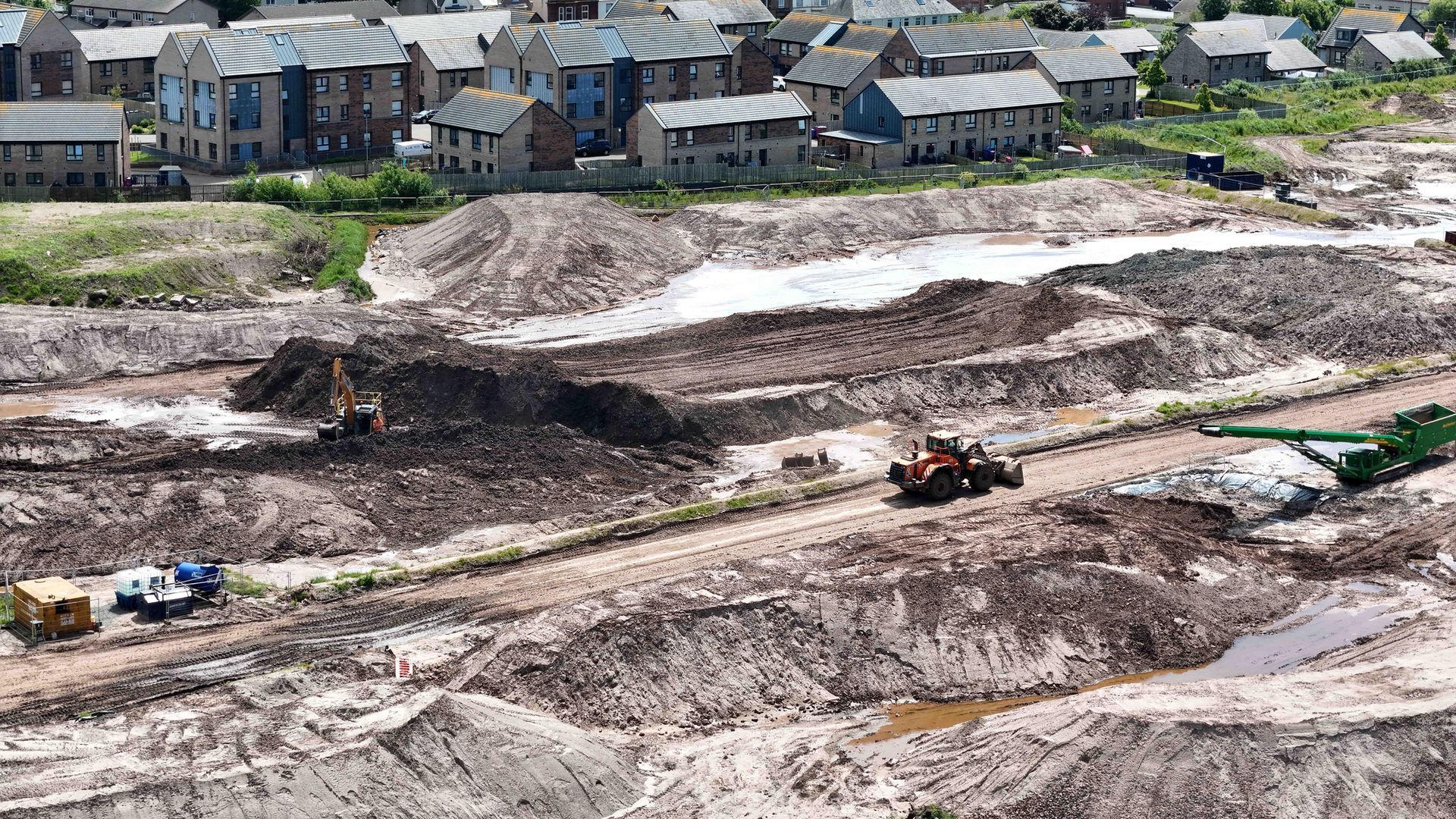 Construction site with heavy machinery, muddy ground, and buildings in the background.