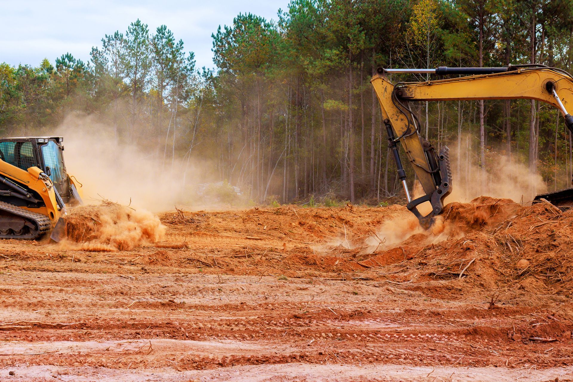 Construction site with excavator and skid steer moving dirt, creating dust.