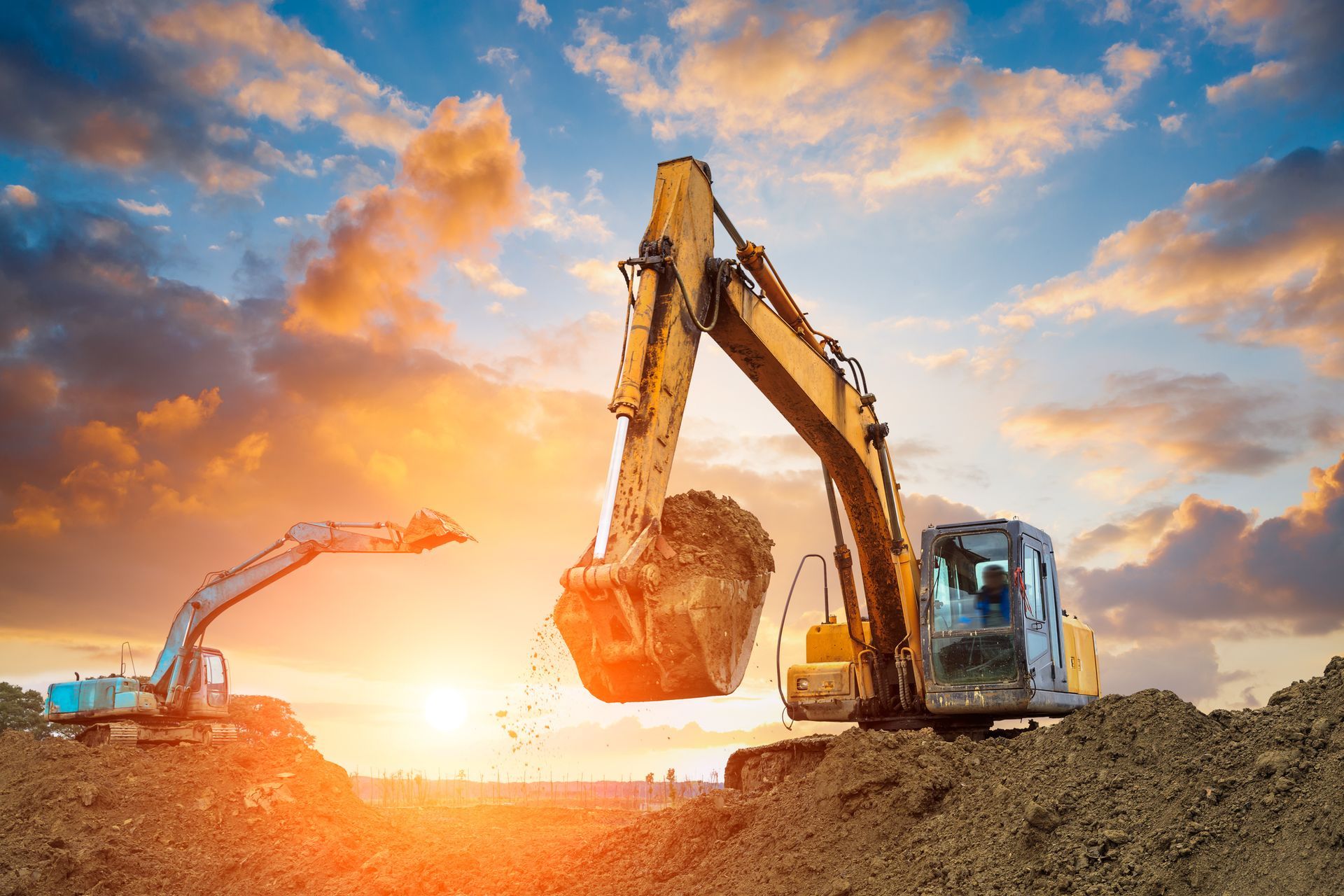 Two excavators at a construction site, scooping dirt against a sunset sky.