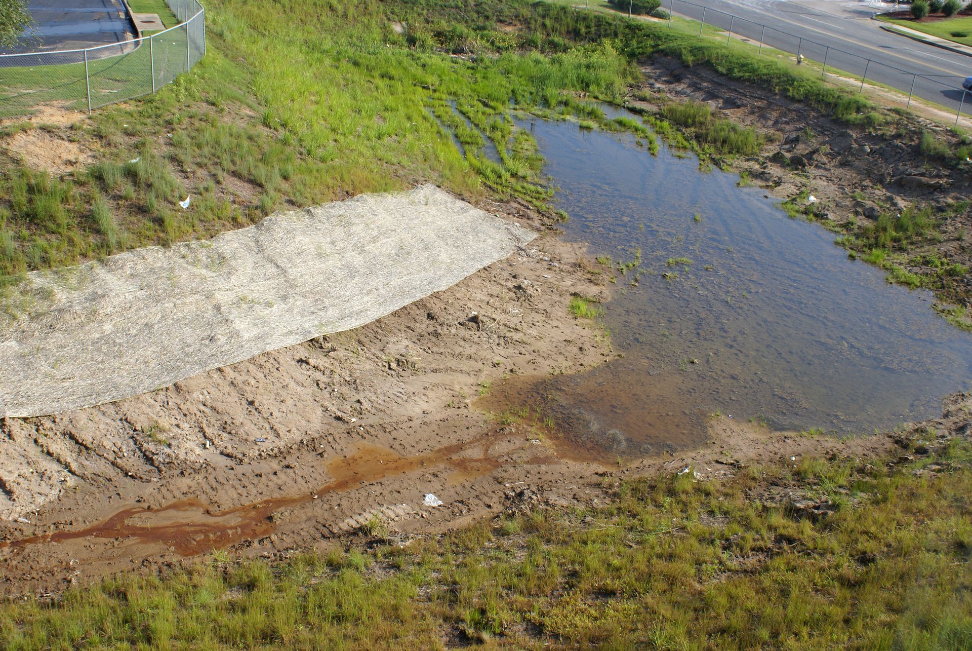 A drainage ditch with pooled water, erosion control materials, and overgrown vegetation near a road.