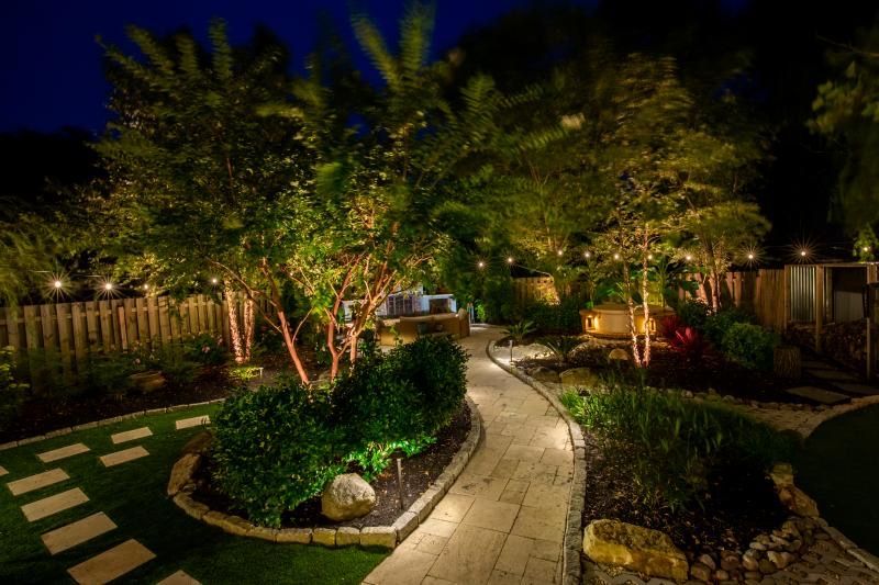 Pathway through a landscaped garden at night, illuminated by soft lighting. Trees, bushes, and a seating area are visible.