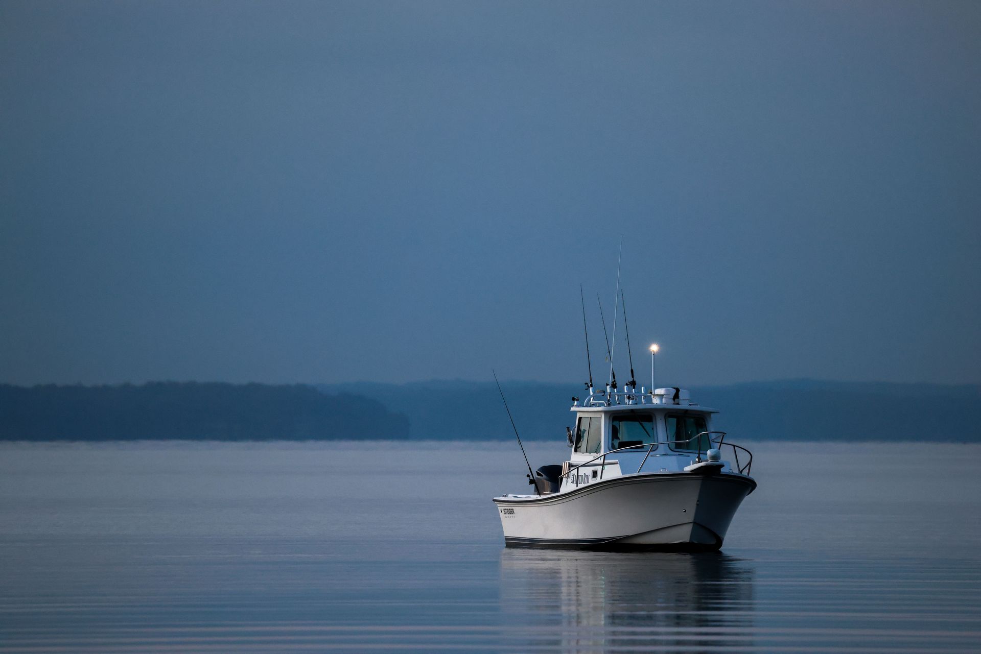 Fishing boat on calm blue water at dusk, centered beneath a hazy horizon.