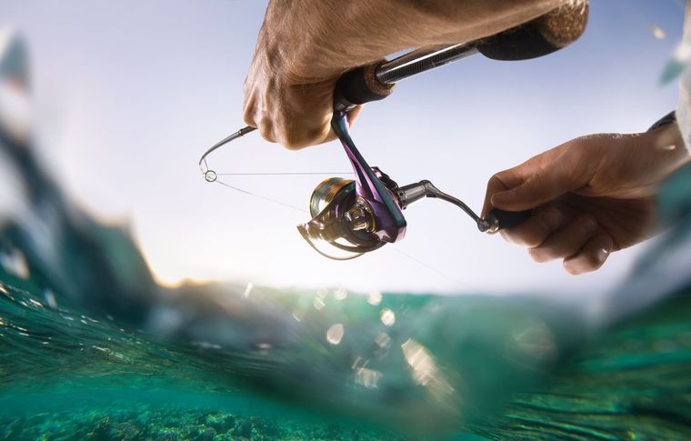 Hands holding a fishing rod above turquoise ocean water, reel and line visible close to the surface