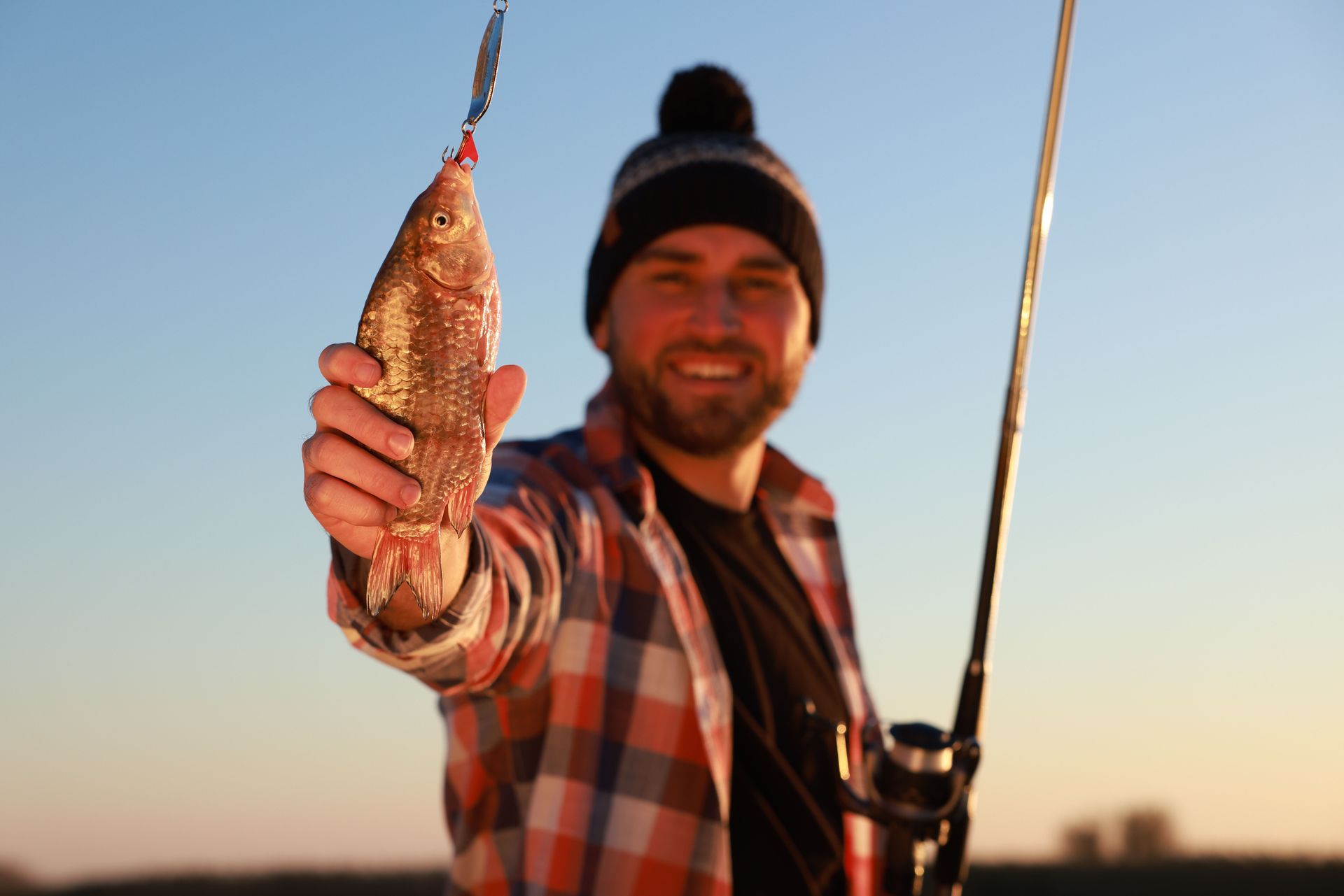 Smiling angler holding a fish by a lake at sunset, with a fishing rod in the background