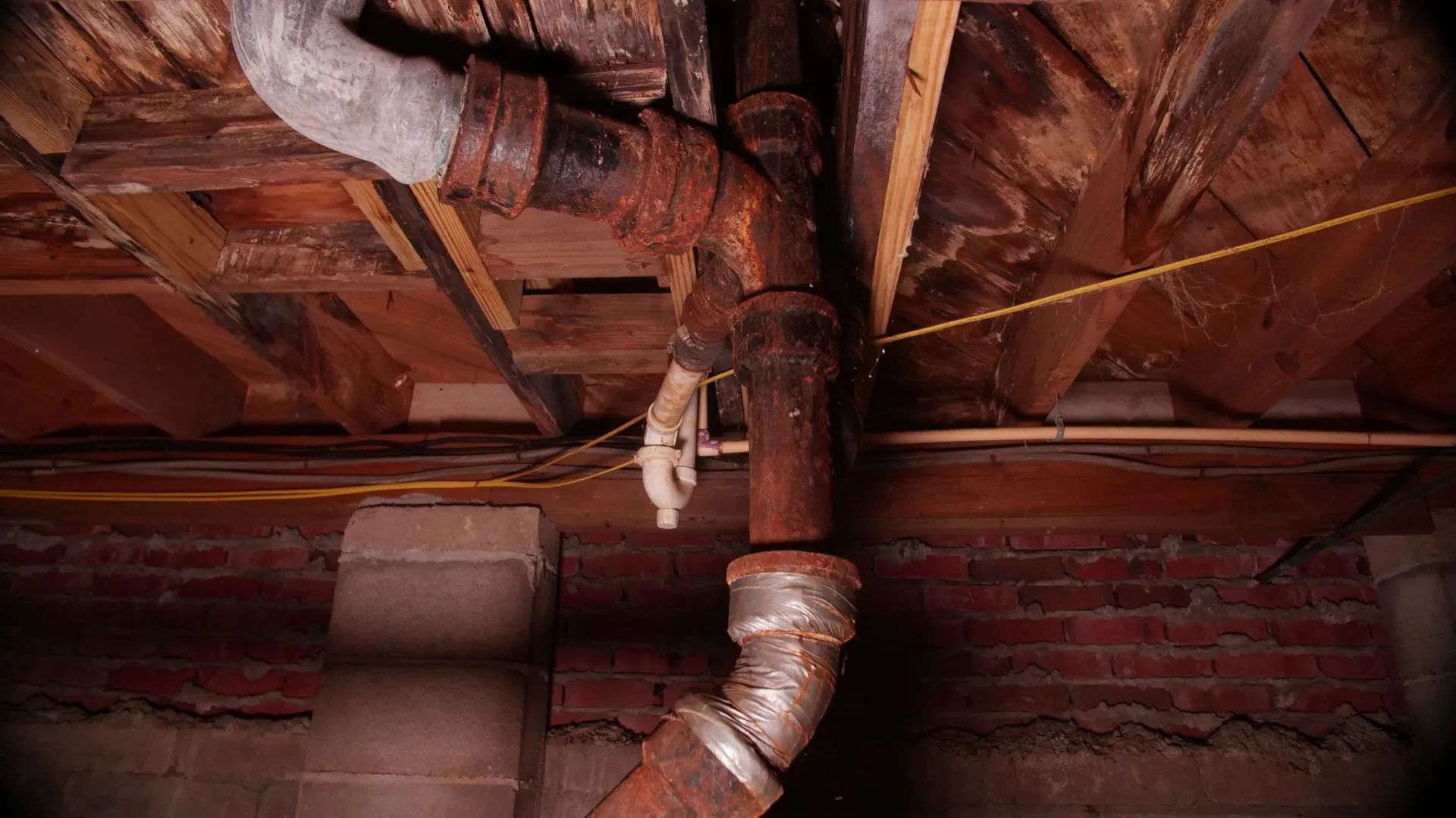Rusting pipes under a wooden ceiling and against a brick wall, likely in a basement.