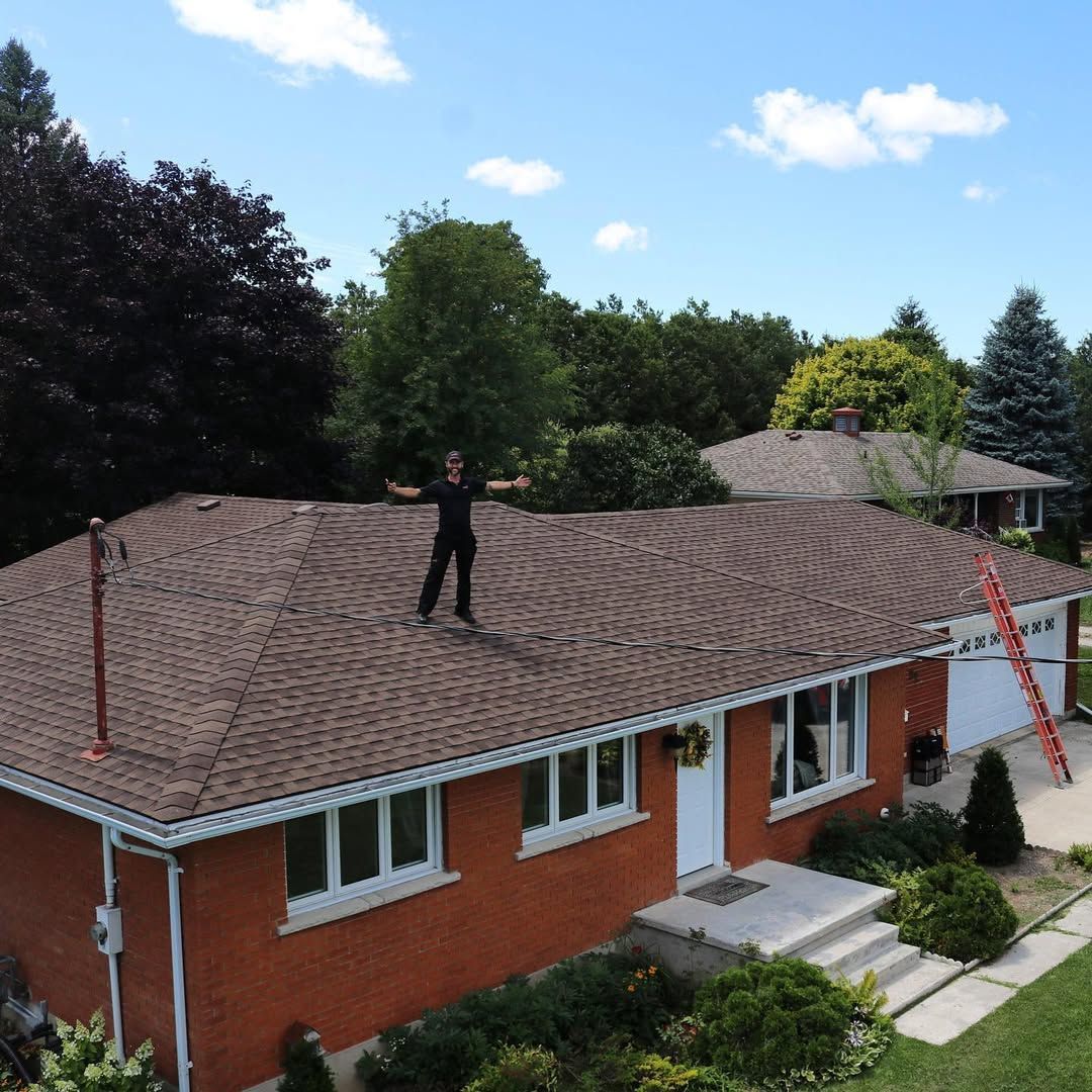 Person standing on a brown-shingled roof of a brick house with trees and a ladder. Blue sky in background.