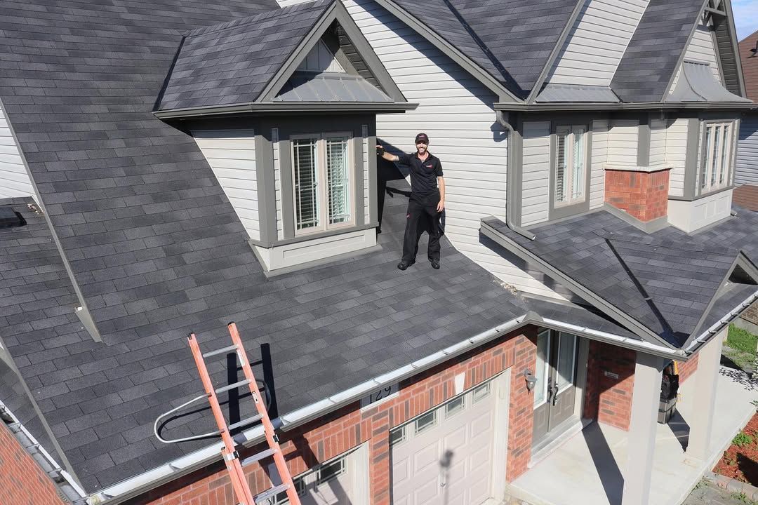 Man in dark clothing stands on a gray shingled roof, near a window. A ladder leans against the roof.