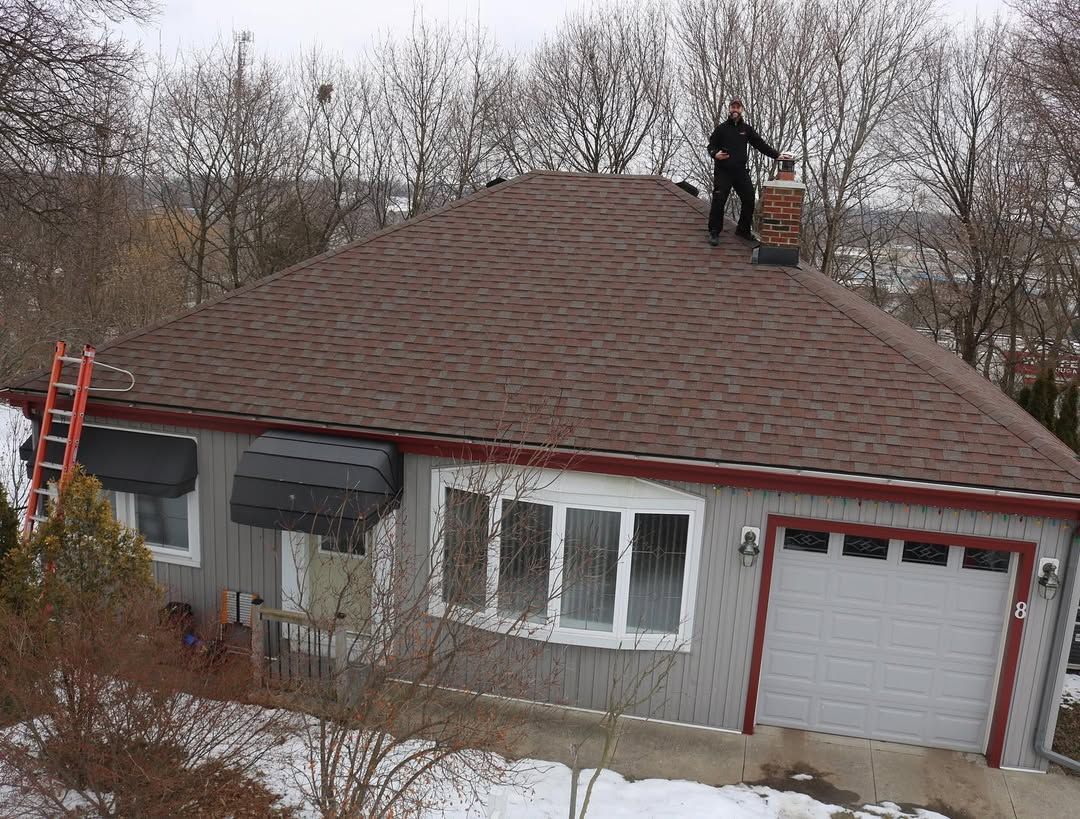 Person on roof, cleaning chimney of a house with brown shingles and a gray exterior. Ladder visible.
