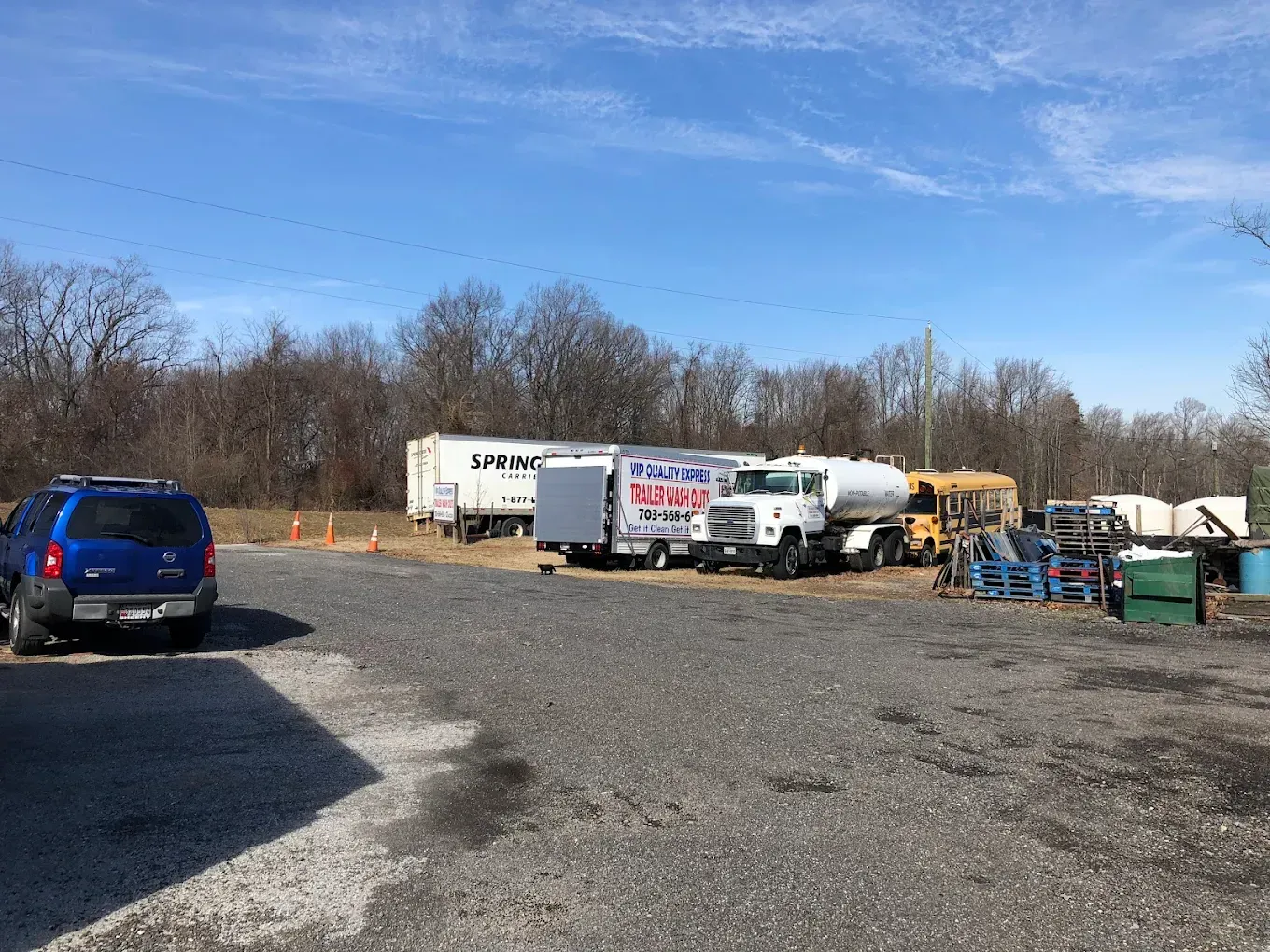Blue SUV parked on gravel with trucks and trailers; sunny day, trees in background.