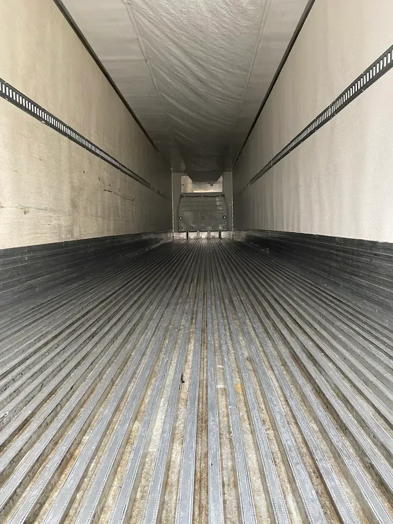 Empty refrigerated semi-trailer interior. Gray, ribbed floor extends toward a distant metal bulkhead.