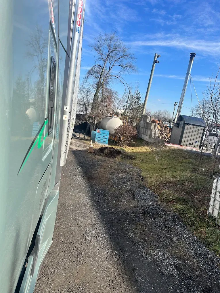 A light-green semi-truck next to a gravel path and grassy area with a bare tree and industrial equipment under a blue sky.