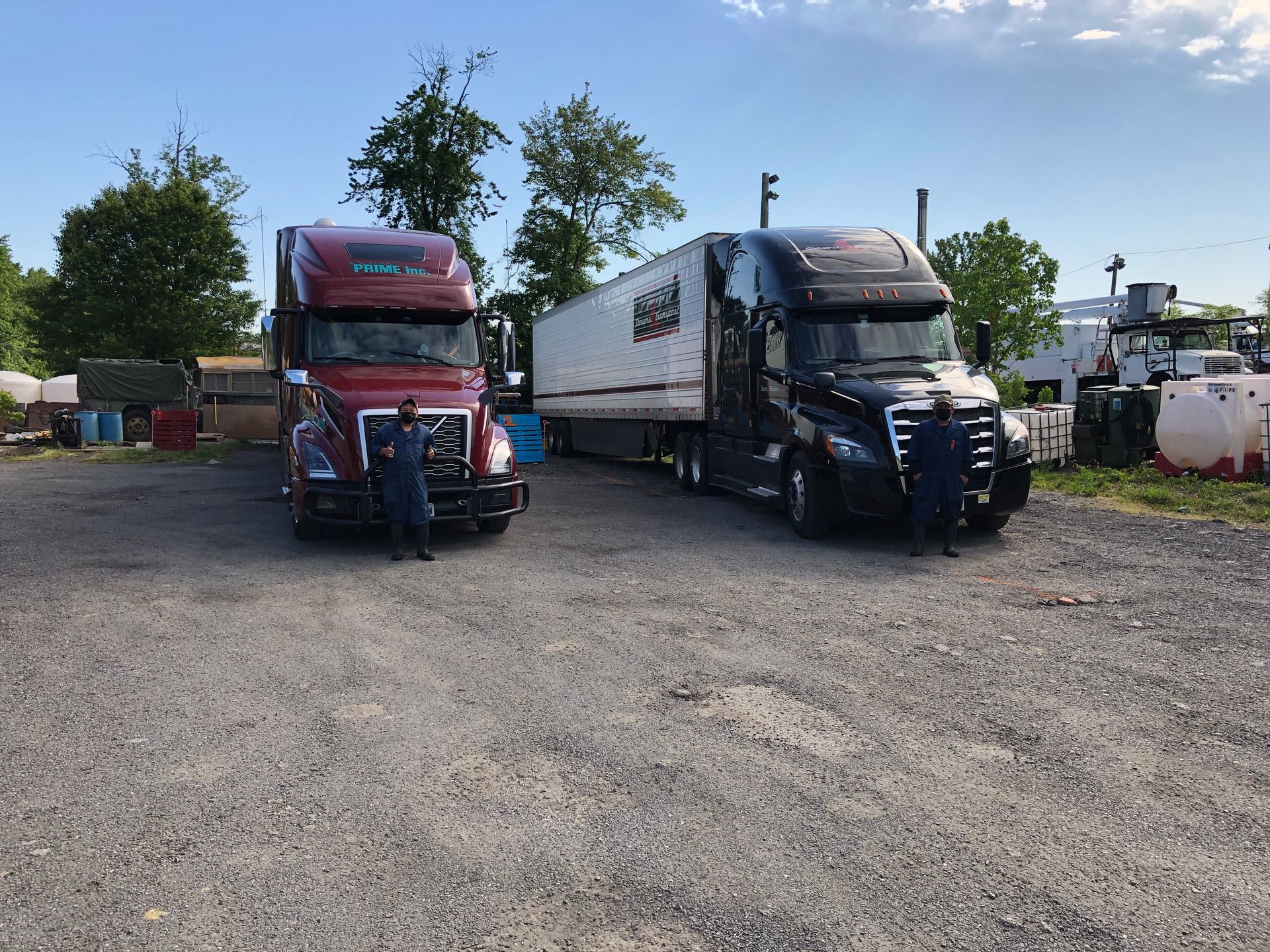 Two semi-trucks parked on a gravel lot; one red, one black with a trailer. People stand in front of the trucks.