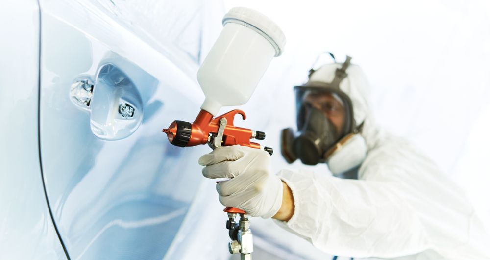 A man wearing a gas mask is spray painting a car.