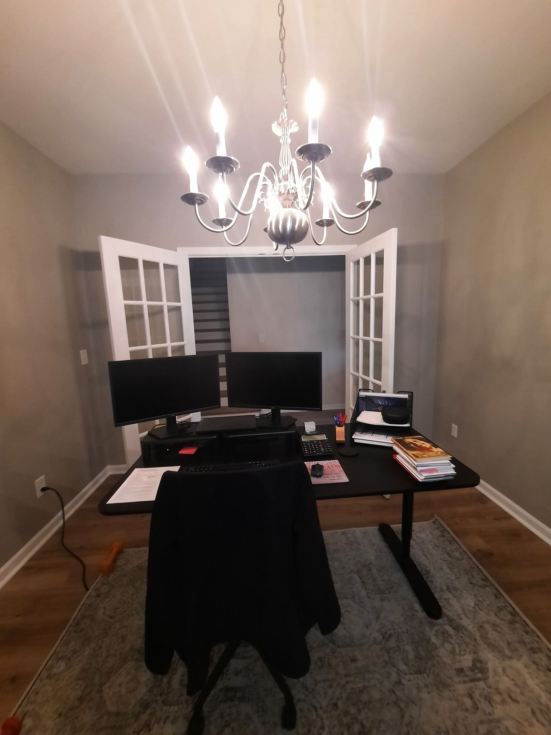 A room with a desk , chair , computer and a chandelier.