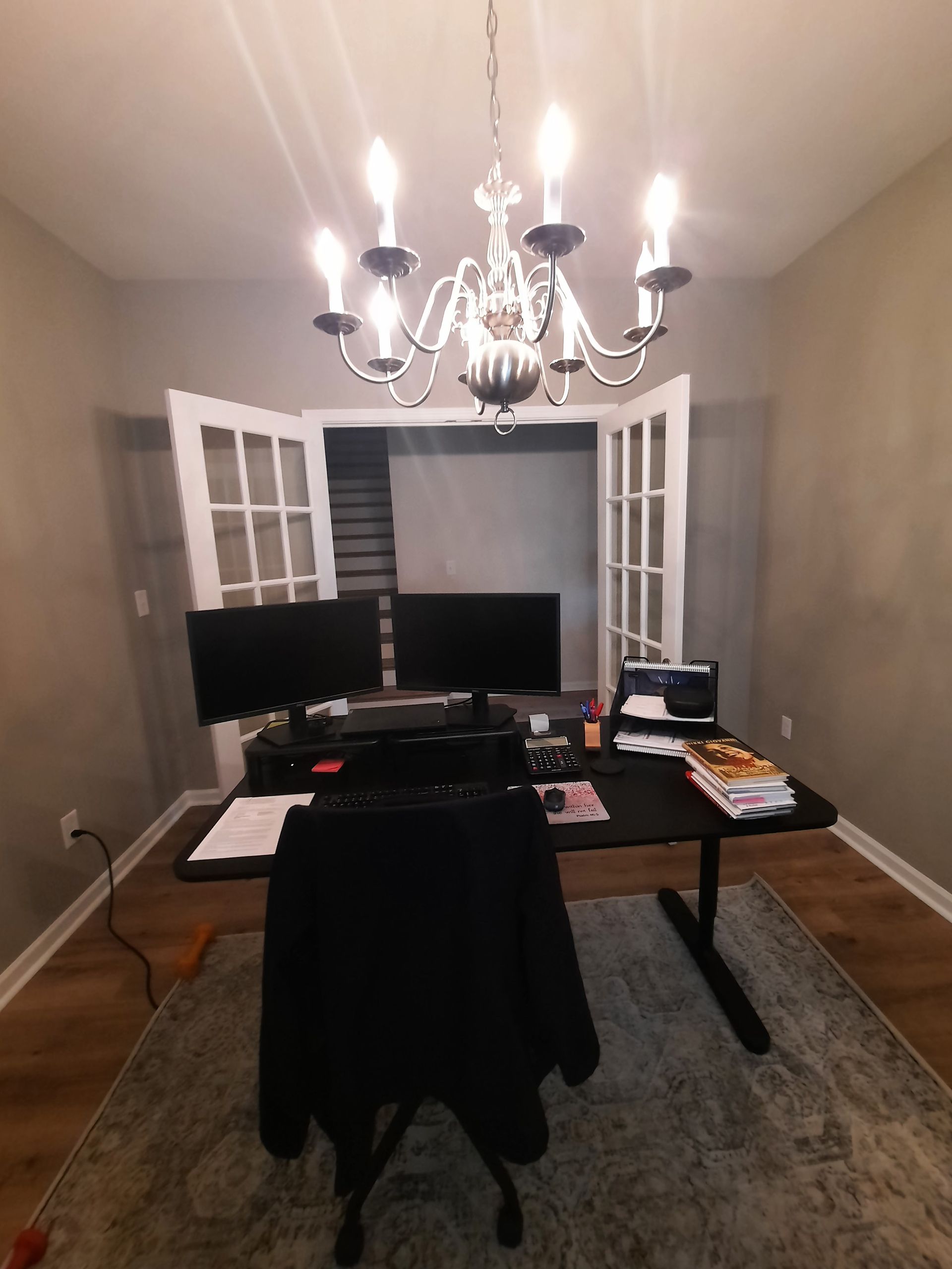 A room with a desk , chair , computer and a chandelier.