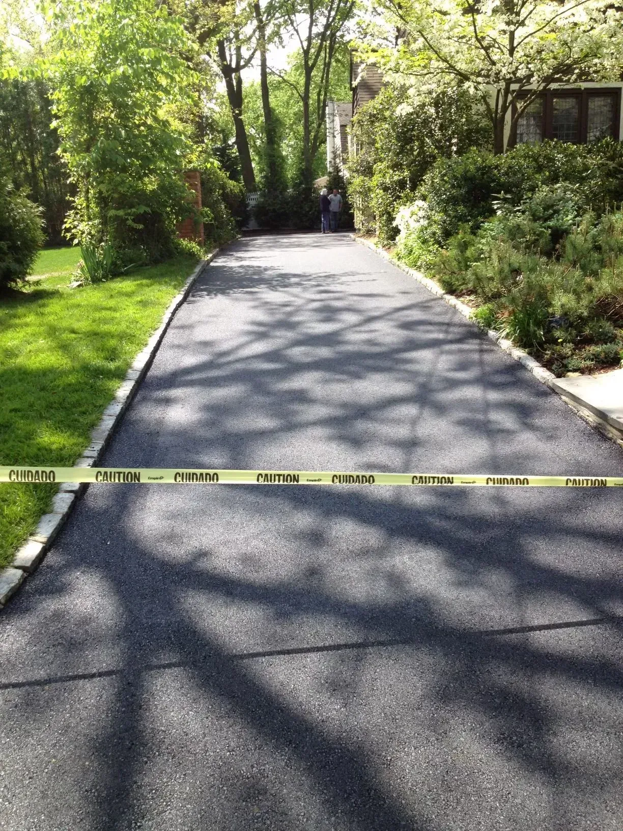 Asphalt driveway blocked by yellow caution tape, bordered by green grass and shrubbery. 