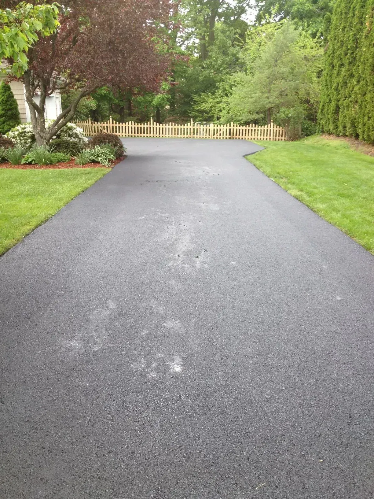 A freshly paved asphalt driveway leads towards a wooden fence and trees, flanked by green grass.