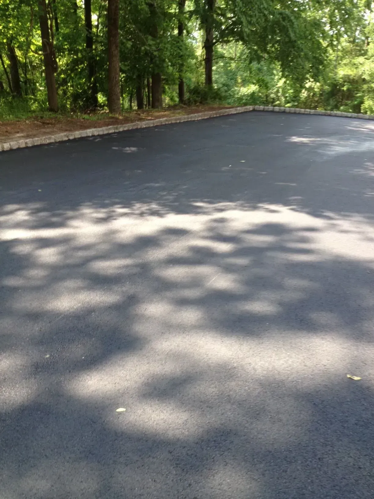 Newly paved asphalt surface with a stone border, shaded by trees in a wooded area.