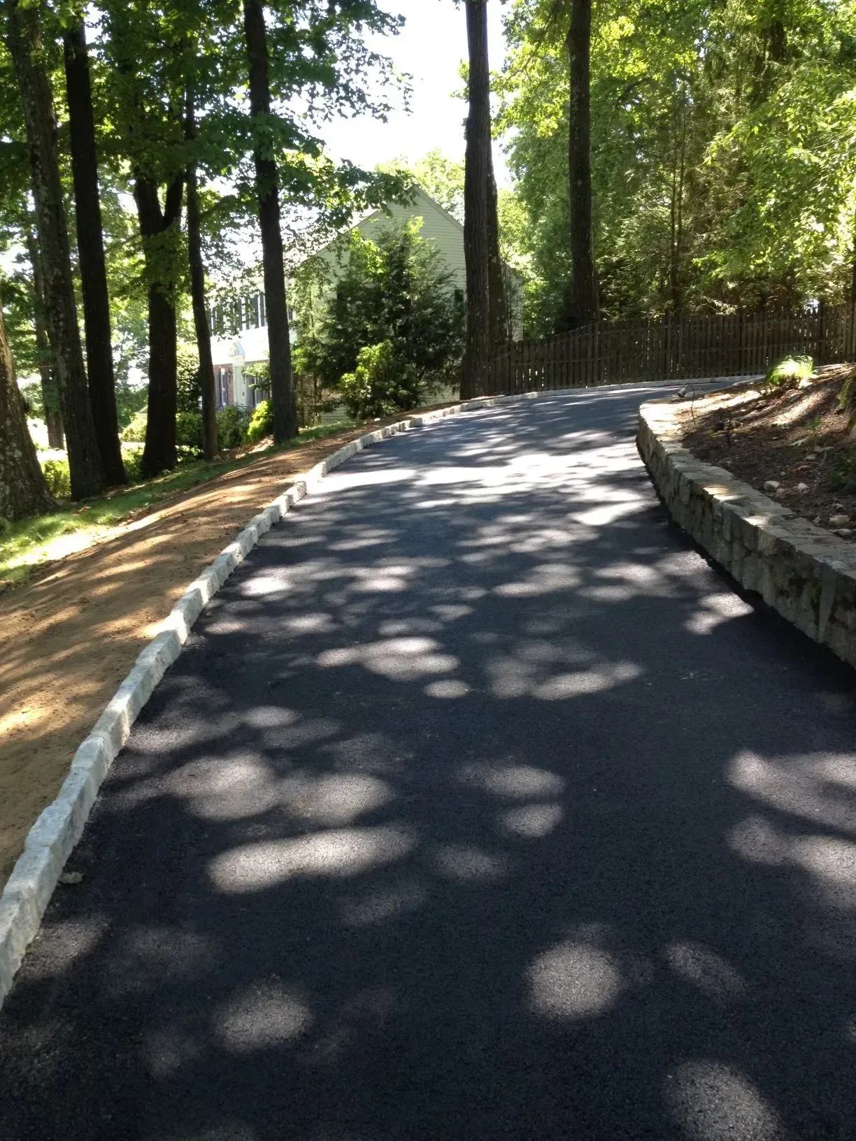 Asphalt driveway winding uphill through a wooded area with stone edging and tree shadows.