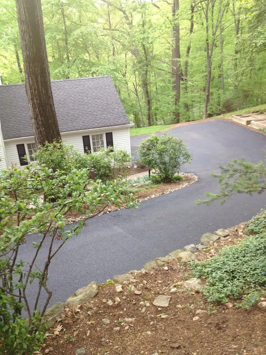 Asphalt driveway curves past a white house with black shutters, through a wooded area with green trees.