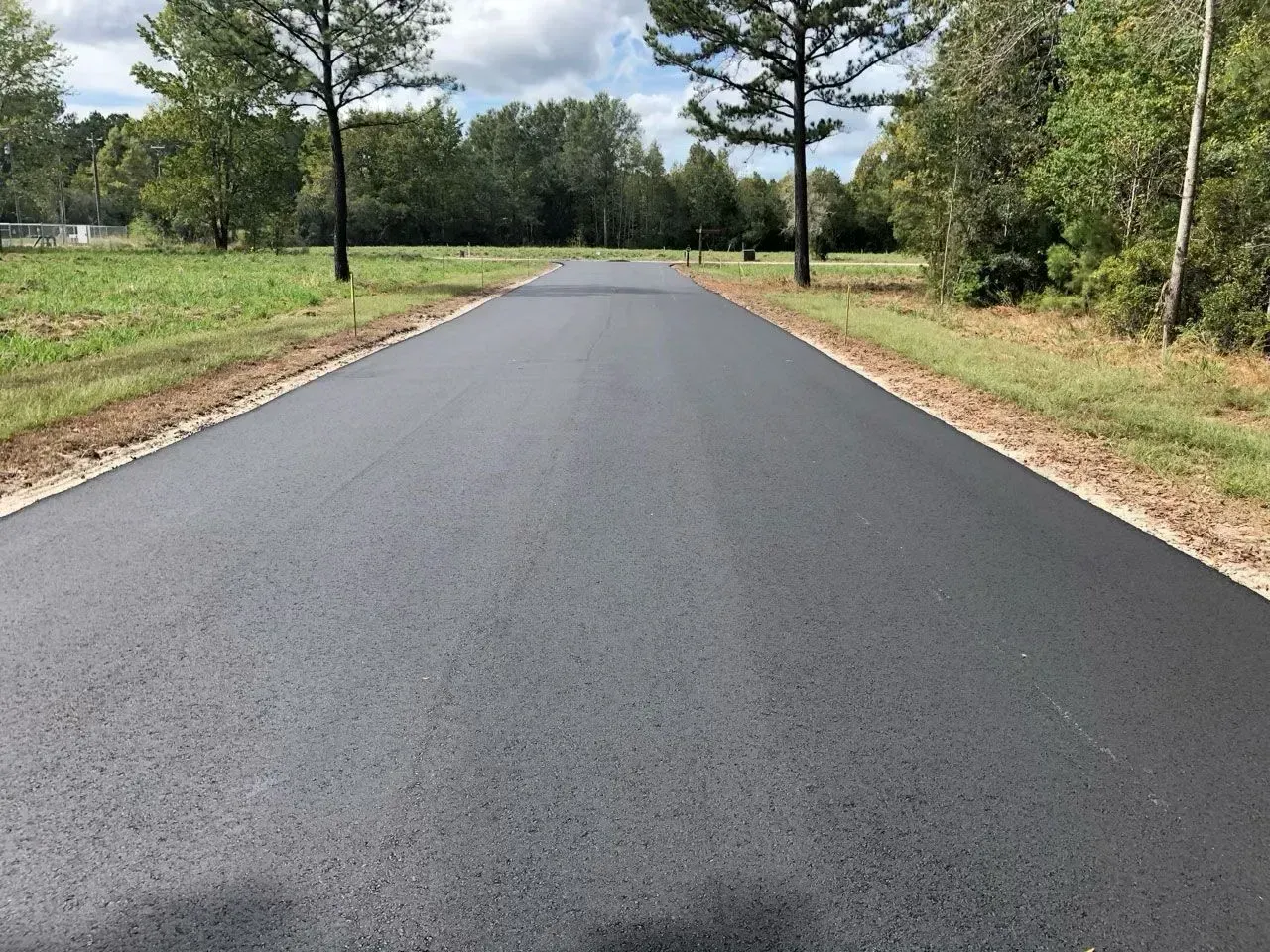 Newly paved asphalt road stretching into the distance, flanked by grassy shoulders and trees under a cloudy sky.
