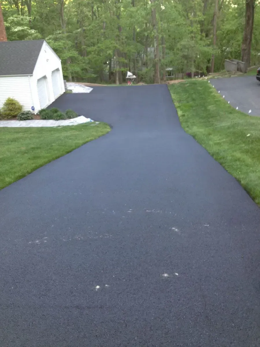 Freshly paved black asphalt driveway curves past a garage and grassy lawn, leading to a tree-lined background.