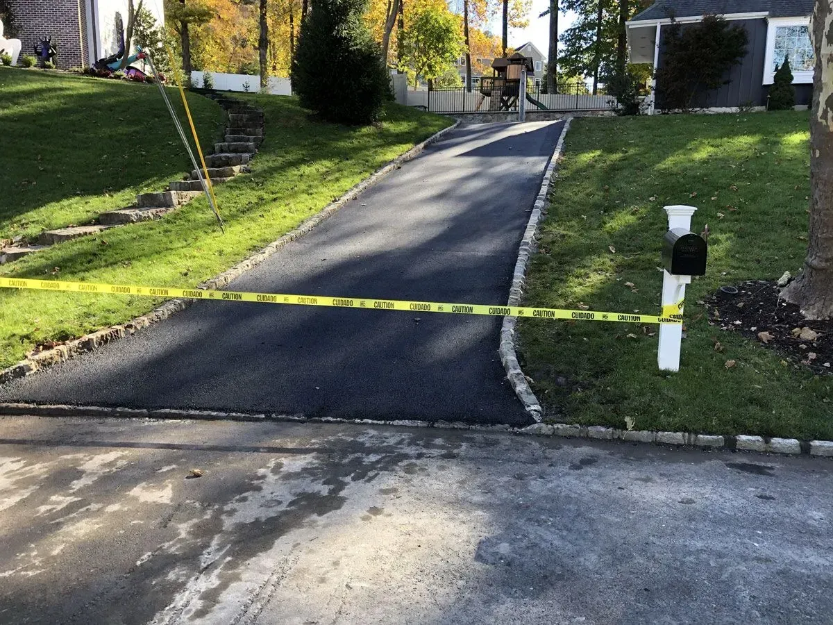 Freshly paved driveway blocked by yellow caution tape, flanked by green grass and a white mailbox.