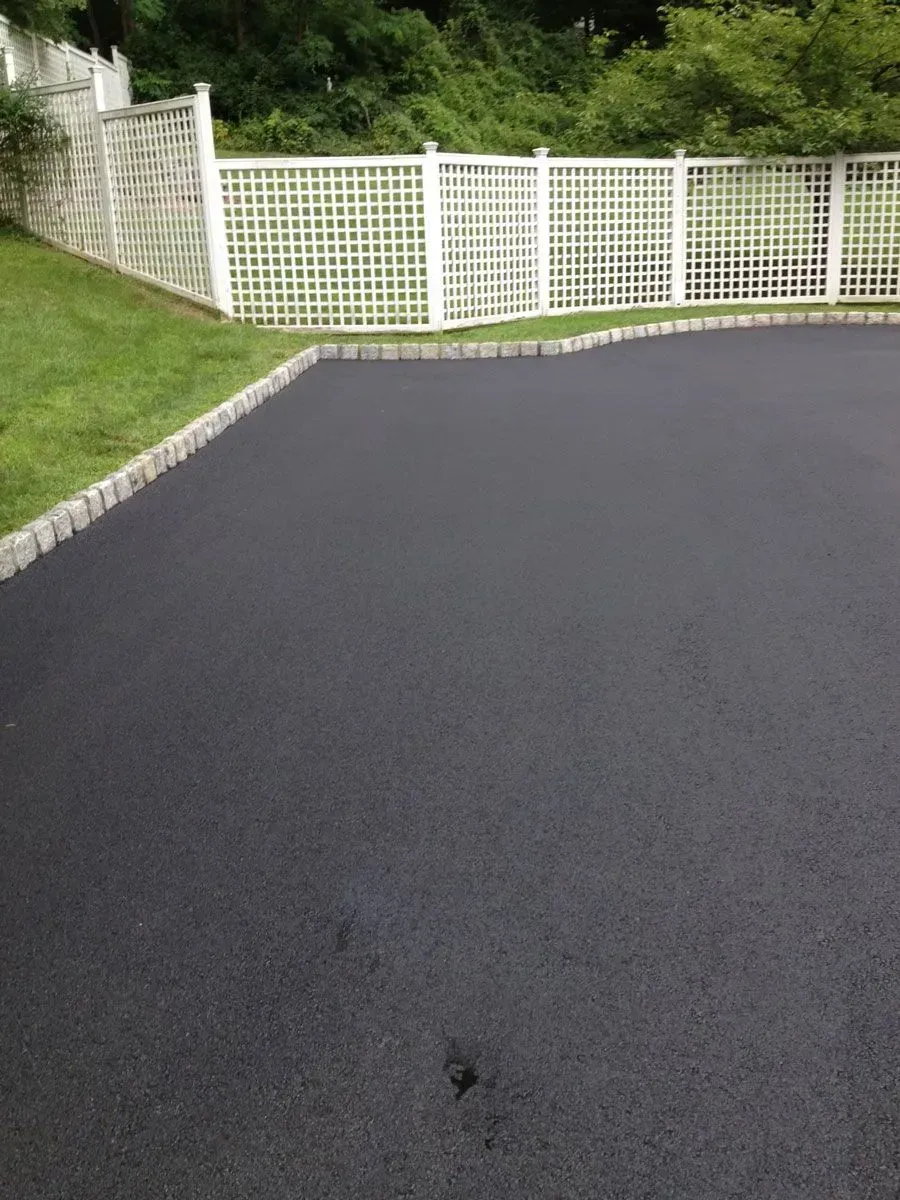 Black asphalt driveway bordered by gray stones and a white latticework fence, set against a green lawn and wooded backdrop.