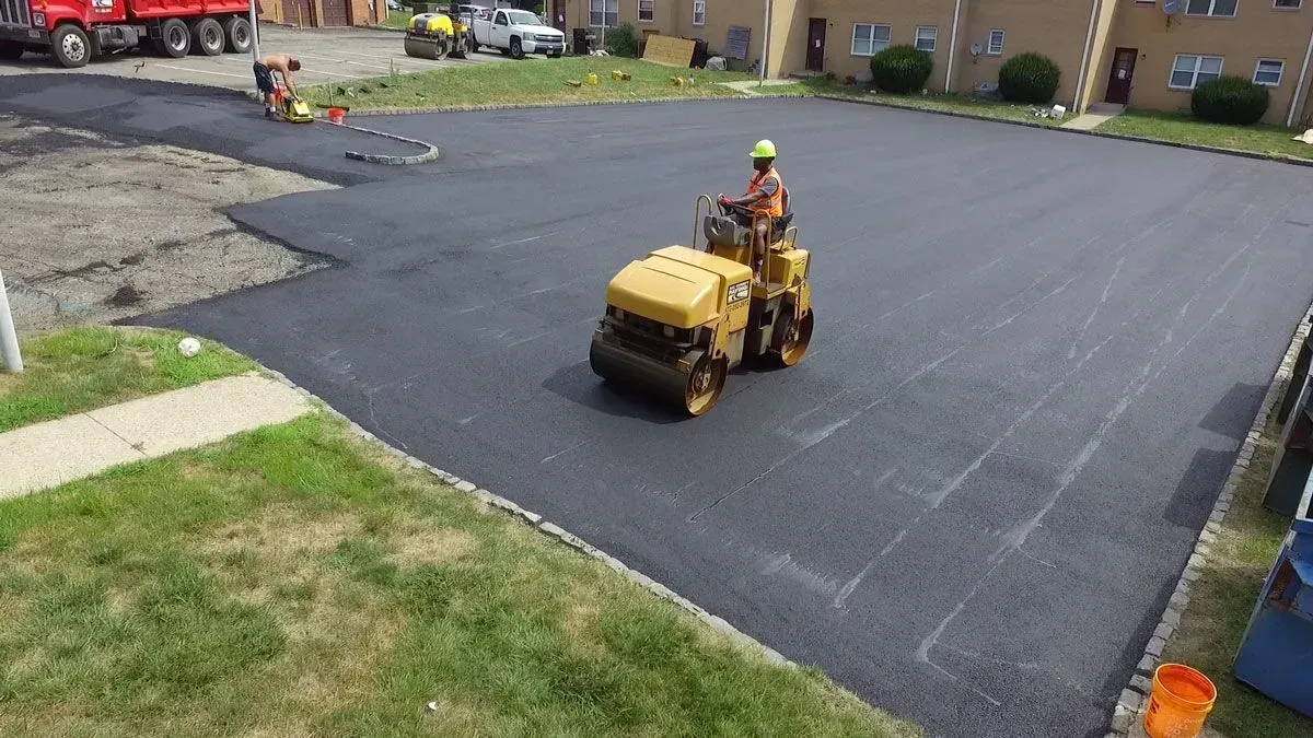 A worker operating a yellow roller compacting fresh asphalt in a parking lot.