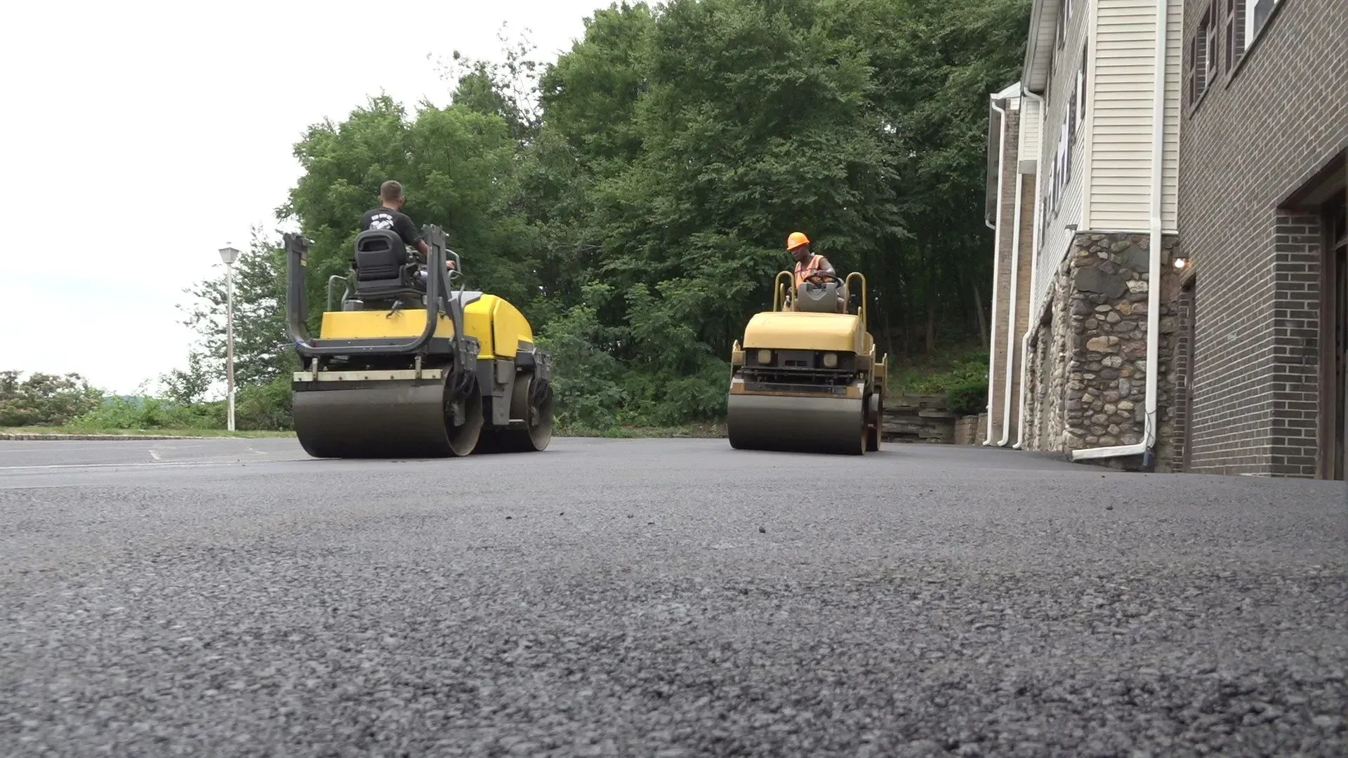 Two yellow asphalt rollers compacting fresh asphalt on a driveway, near a building and trees.