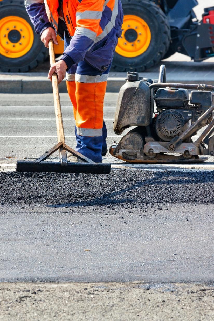 Person in reflective workwear raking asphalt on a road, with a compactor and construction equipment visible.