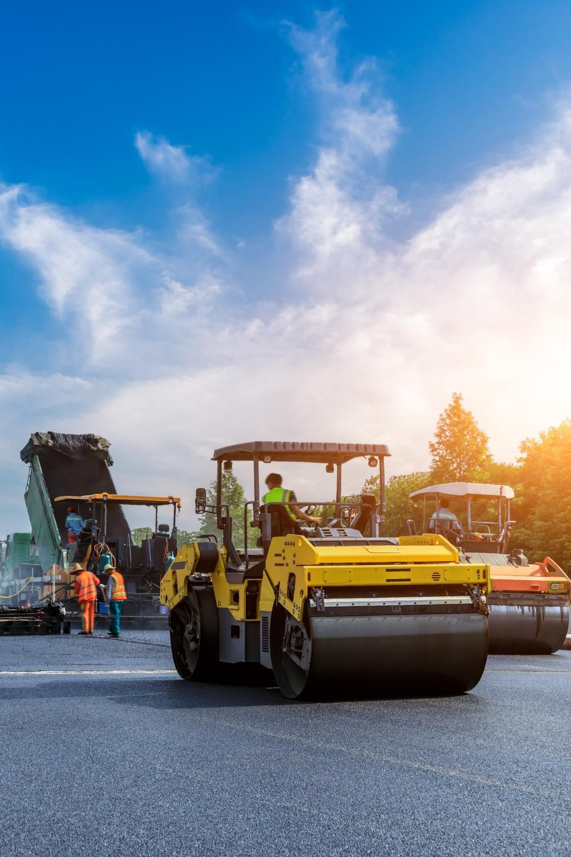 Road construction scene with a yellow steamroller compacting freshly laid asphalt under a blue sky. 