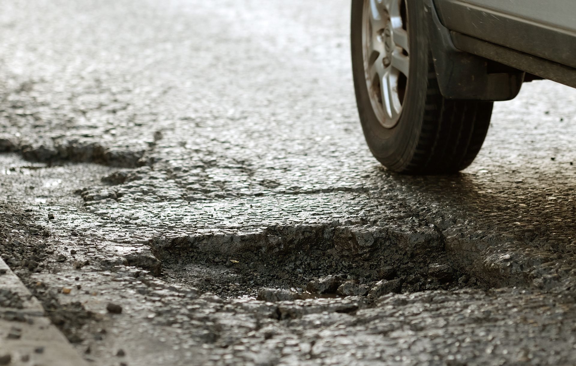 Car tire near a pothole on a worn, cracked asphalt road.
