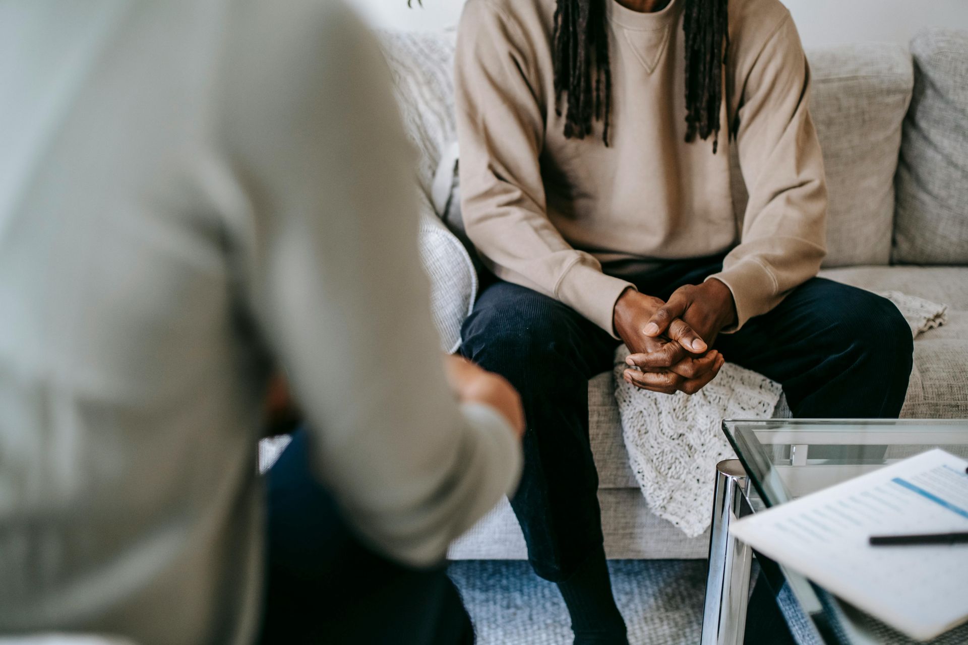 Two people in a therapy session, one seated on a couch with hands clasped across from a blurred listener.