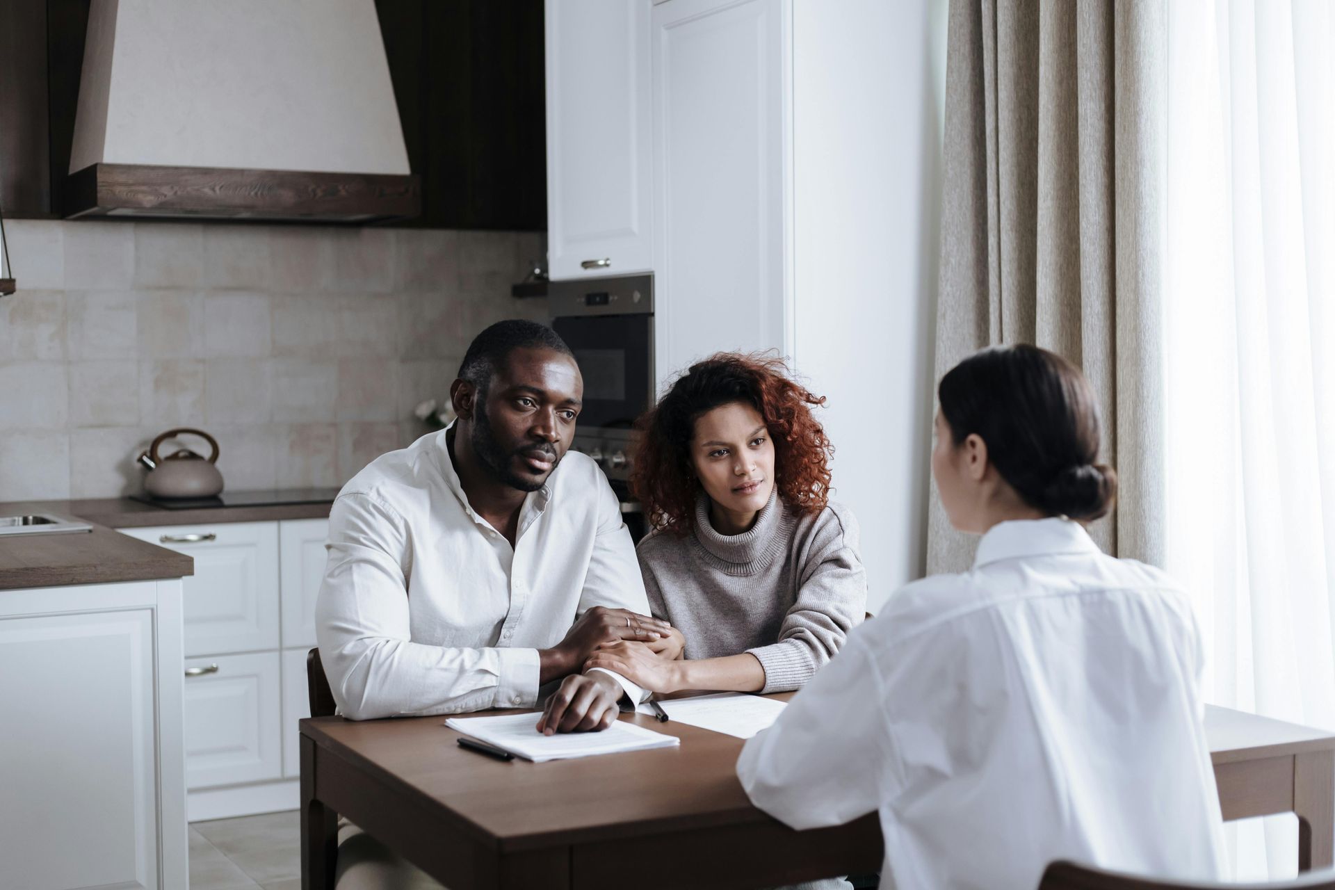 Three people discussing paperwork at a kitchen table with a laptop in a bright home interior