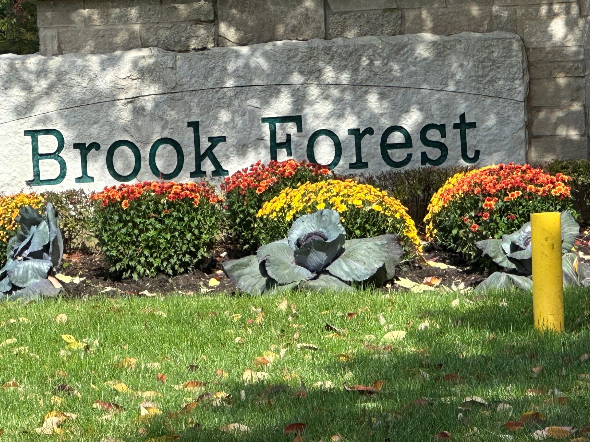 A sign for brook forest is surrounded by flowers and a yellow pole