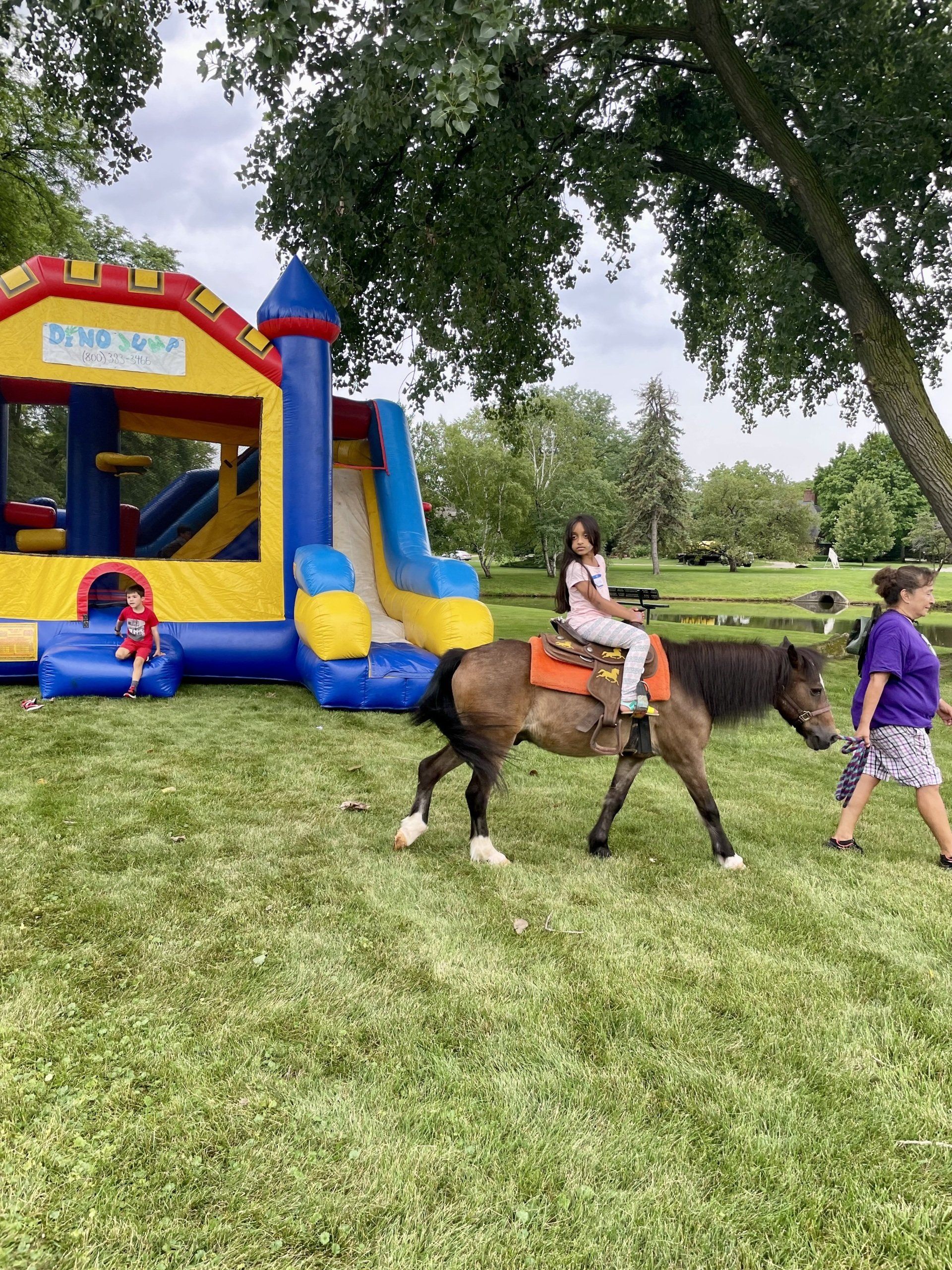 A little girl is riding a pony in front of a bouncy house.